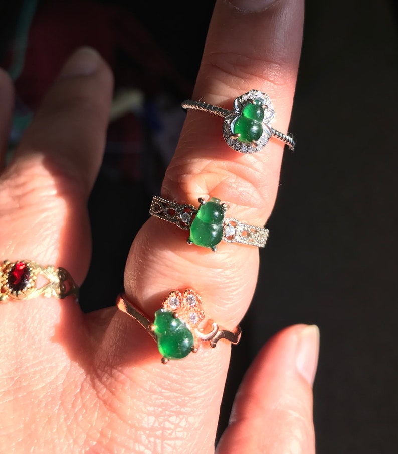 May include: Three jade rings with silver and gold settings. The jade stones are green and shaped like gourds. The rings are on a finger.