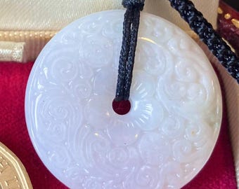 Op de afbeelding: Een witte jade hanger met een gegraveerd bloemenmotief, hangend aan een zwart koord.