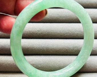 Pode incluir: Uma pulseira de jade verde claro. A pulseira circular é de cor verde translúcida sólida. O jade tem uma superfície lisa e polida. A pulseira é segurada por uma mão sobre um fundo cinza.