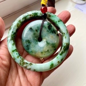 May include: A green jade pendant with a ring design. The pendant is on a brown cord with red and yellow beads.