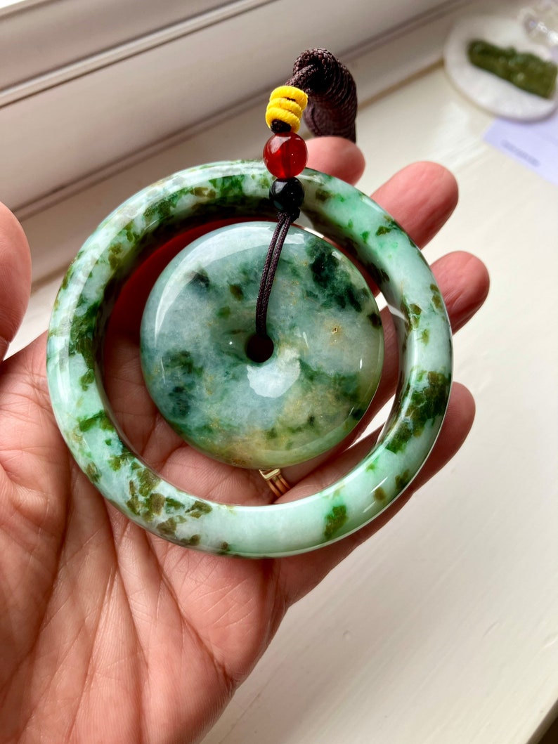 May include: A green jade pendant with a ring design. The pendant is on a brown cord with red and yellow beads.