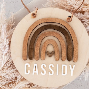 May include: A wooden round sign with a brown rainbow design and the name "CASSIDY" in white letters. The sign has a brown leather cord for hanging.