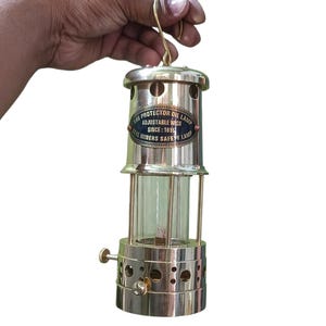 May include: A vintage brass oil lamp with a glass cylinder and a handle. The lamp has the text "THE PROTECTOR OIL LAMP ADJUSTABLE WICK SINCE: 1815 COAL MINERS SAFETY LAMP" on a black and gold label. The lamp is held by a hand.