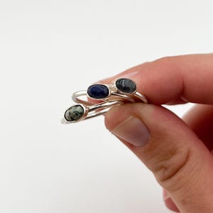 May include: Three silver rings with oval gemstones in shades of blue and green. The rings are held against a white background, showcasing the polished metal and the colorful stones. The gemstones have unique patterns and textures.
