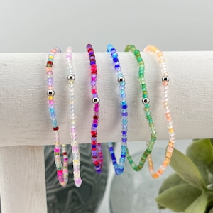 May include: Six colorful beaded bracelets with silver beads. The bracelets are arranged in a row on a white surface.