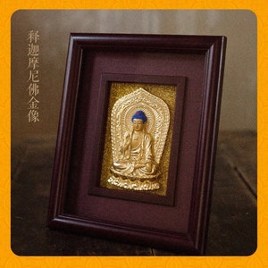 May include: A framed gold-colored Buddha statue with a blue crown, set against a brown background. The frame is dark brown wood with a gold-colored inner border. The text "释迦摩尼佛金像" is written in black on a white background.