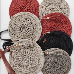 Pu&ograve; includere: Sei borse rotonde all'uncinetto in vari colori, tra cui beige, nero e rosso. Le borse hanno un design circolare con un fiocco appeso a ciascuna. Una borsa ha una tracolla in pelle marrone.