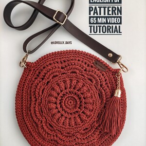 Pu&ograve; includere: Una borsa rotonda di uncinetto rossa con una tracolla in pelle marrone e un fiocco dorato. La borsa ha un motivo circolare con un design a mandala. L'immagine include il testo "ENGLISH PDF PATTERN 65 MIN VIDEO TUTORIAL @LOVELLY_DAYS".