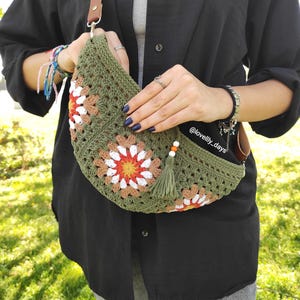 May include: Olive green crochet bag with a brown leather strap. The bag features floral patterns in white, orange, and tan. A green tassel and wooden beads hang from the side. The bag is held by a person wearing a black shirt and jeans.