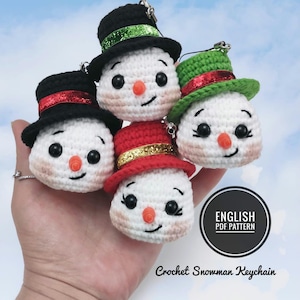 Op de afbeelding: Vier gehaakte sneeuwpop sleutelhangers met verschillende gekleurde hoeden. De sneeuwpoppen hebben zwarte ogen, rode neuzen en zwarte monden. De hoeden zijn zwart met rode, groene of gouden versieringen. De tekst "ENGLISH PDF PATTERN" en "Crochet Snowman Keychain" is zichtbaar onder de sneeuwpoppen.