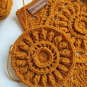 May include: A close-up of a crocheted mustard yellow mandala with a smaller circle in the center. The mandala is made with a fuzzy yarn and has a hand-made tag attached.