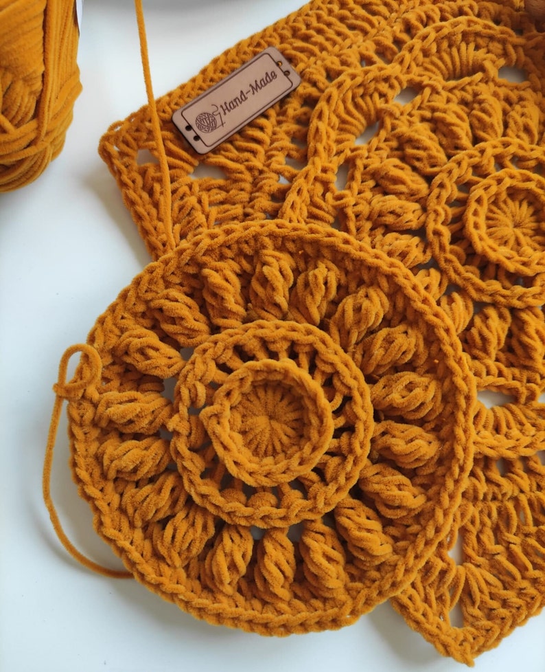 May include: A close-up of a crocheted mustard yellow mandala with a smaller circle in the center. The mandala is made with a fuzzy yarn and has a hand-made tag attached.