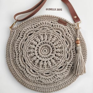 Puede incluir: Un bolso redondo de crochet beige con una correa de cuero marr&oacute;n y un borla. El bolso tiene un dise&ntilde;o circular con un patr&oacute;n de mandala. La correa tiene una hebilla dorada y el bolso tiene un dije de cuentas de madera.