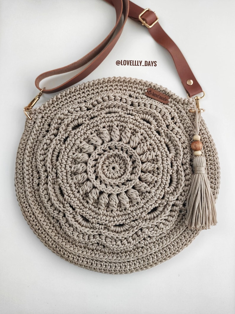 Puede incluir: Un bolso redondo de crochet beige con una correa de cuero marr&oacute;n y un borla. El bolso tiene un dise&ntilde;o circular con un patr&oacute;n de mandala. La correa tiene una hebilla dorada y el bolso tiene un dije de cuentas de madera.