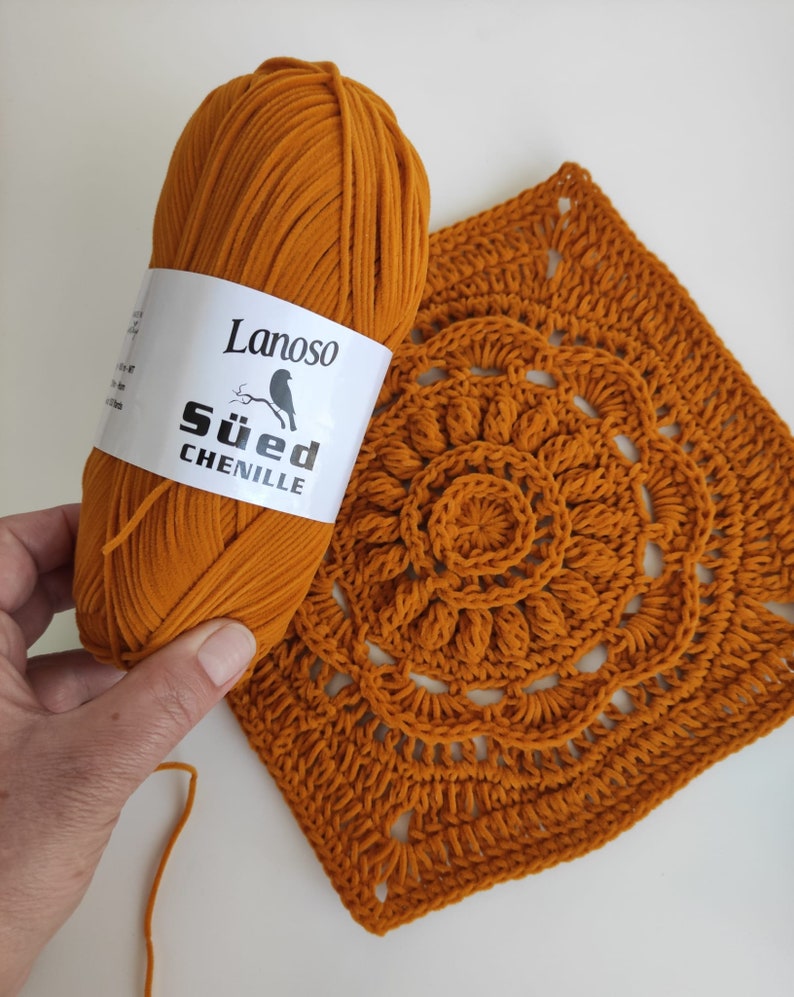 May include: A ball of orange yarn labeled "Lanoso S&uuml;d Chenille" next to a crocheted square with a circular design in the center.