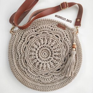 Pu&ograve; includere: Una borsa a tracolla rotonda in crochet beige con una tracolla in pelle marrone e un pendente a forma di nappa con perline di legno. La borsa presenta un design circolare con un motivo a mandala. La tracolla ha una fibbia dorata e il testo "@LOVELLY_DAYS" &egrave; visibile sulla borsa.