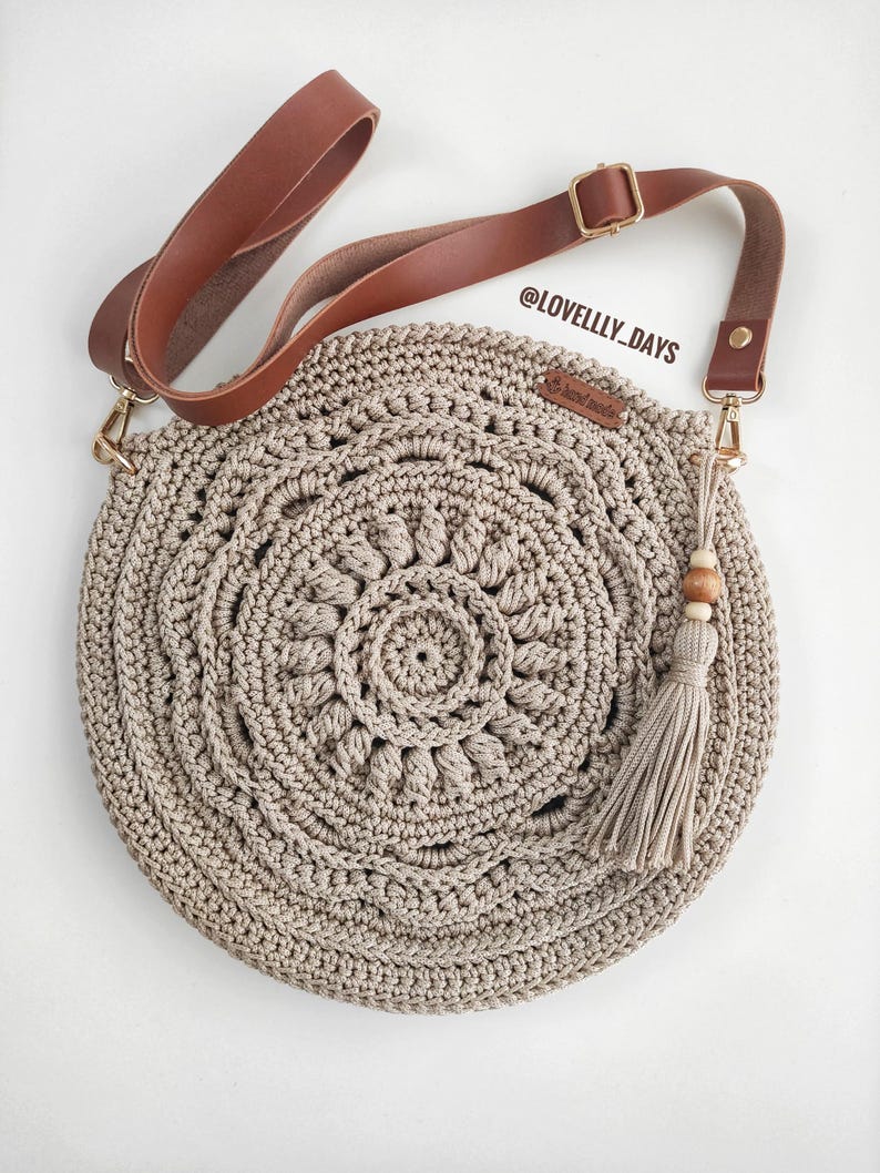 Pu&ograve; includere: Una borsa a tracolla rotonda in crochet beige con una tracolla in pelle marrone e un pendente a forma di nappa con perline di legno. La borsa presenta un design circolare con un motivo a mandala. La tracolla ha una fibbia dorata e il testo "@LOVELLY_DAYS" &egrave; visibile sulla borsa.