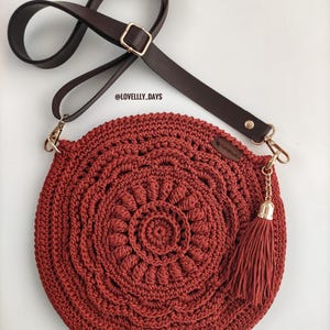 Pu&ograve; includere: Una borsa a mano rotonda in crochet con una tracolla in pelle marrone e un fiocco dorato. La borsa &egrave; di colore rosso scuro e presenta un motivo circolare.
