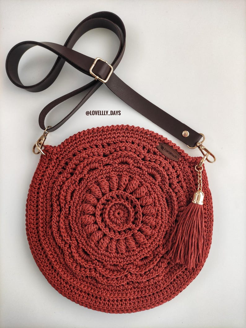 Pu&ograve; includere: Una borsa a mano rotonda in crochet con una tracolla in pelle marrone e un fiocco dorato. La borsa &egrave; di colore rosso scuro e presenta un motivo circolare.