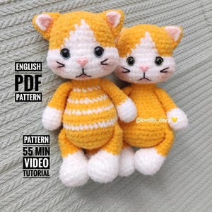 May include: Two crocheted cat toys, one orange and white and one yellow and white. The larger cat is wearing a striped sweater. The smaller cat is sitting behind the larger cat. The text "ENGLISH PDF PATTERN" and "PATTERN 55 MIN VIDEO TUTORIAL" is visible on the image. The text "@lovelily_days" is also visible on the image.