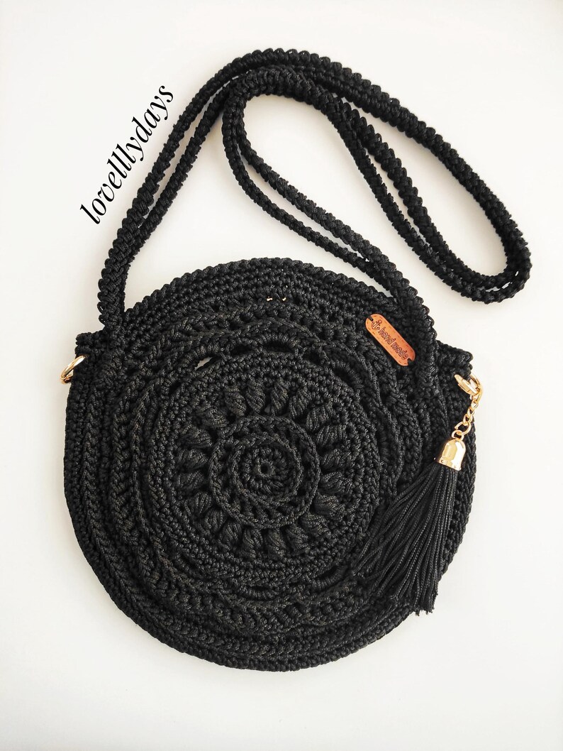 Pu&ograve; includere: Borsa a tracolla rotonda in crochet nero con una catena dorata e un pon pon. La borsa ha un piccolo cartellino in pelle marrone con il testo "lovelydays" su di esso.