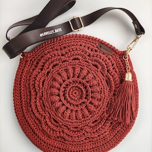 Puede incluir: Un bolso redondo de crochet rojo con una correa de cuero marr&oacute;n y una cadena dorada con un borla. El bolso tiene un dise&ntilde;o circular con un c&iacute;rculo m&aacute;s peque&ntilde;o en el centro. El bolso tiene una etiqueta que dice "Handmade".