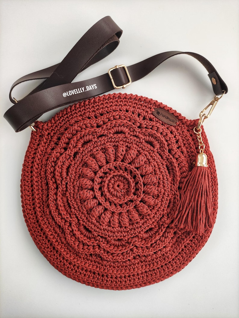 Puede incluir: Un bolso redondo de crochet rojo con una correa de cuero marr&oacute;n y una cadena dorada con un borla. El bolso tiene un dise&ntilde;o circular con un c&iacute;rculo m&aacute;s peque&ntilde;o en el centro. El bolso tiene una etiqueta que dice "Handmade".