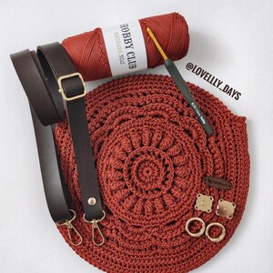 Pu&ograve; includere: Una borsa all'uncinetto di colore ruggine con una tracolla in pelle marrone e accessori dorati. La borsa &egrave; realizzata con un motivo circolare e ha un'etichetta che dice "Hobby Club Macrame No.3".