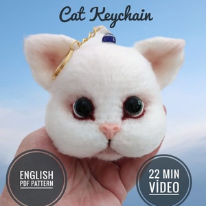 Puede incluir: Llavero de gato de fieltro blanco con un dije de ojo azul y blanco. El llavero está sostenido en una mano. El texto "Cat Keychain" está en la parte superior de la imagen. El texto "ENGLISH PDF PATTERN" está en un círculo en la parte inferior izquierda de la imagen. El texto "22 MIN VIDEO" está en un círculo en la parte inferior derecha de la imagen.