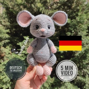Può includere: Un giocattolo di topo di uncinetto grigio con orecchie e piedi rosa. Il topo è seduto in una mano. Lo sfondo include una bandiera tedesca e il testo "5 MIN VIDEO" e "DEUTSCH PDF ANLEITUNG".