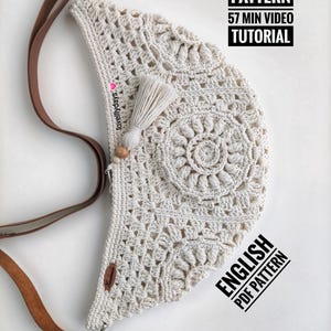 Può includere: Una borsa color crema realizzata a uncinetto con una tracolla in pelle marrone. La borsa ha un fiocco e una perla di legno. Il testo "PATTERN 57 MIN VIDEO TUTORIAL" e "ENGLISH PDF PATTERN" è visibile nell'immagine.