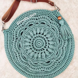 Puede incluir: Un bolso de crochet de color turquesa con una correa de cuero marr&oacute;n y un borla de cuentas de madera. El bolso tiene forma circular y un dise&ntilde;o de mandala. El texto "@lovelily_days" es visible en el bolso.