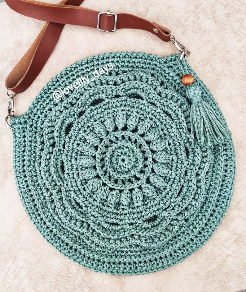 Puede incluir: Un bolso de crochet de color turquesa con una correa de cuero marr&oacute;n y un borla de cuentas de madera. El bolso tiene forma circular y un dise&ntilde;o de mandala. El texto "@lovelily_days" es visible en el bolso.