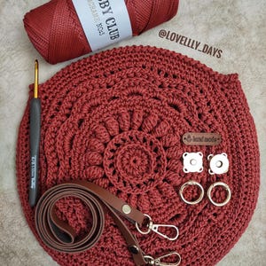 Pu&ograve; includere: Una borsa a uncinetto rossa con una tracolla in pelle marrone e accessori dorati. La borsa &egrave; realizzata con un cordoncino rosso etichettato "Hobby Club Macrame No.3". La borsa ha anche un piccolo cartellino "fatto a mano" e due chiusure magnetiche.
