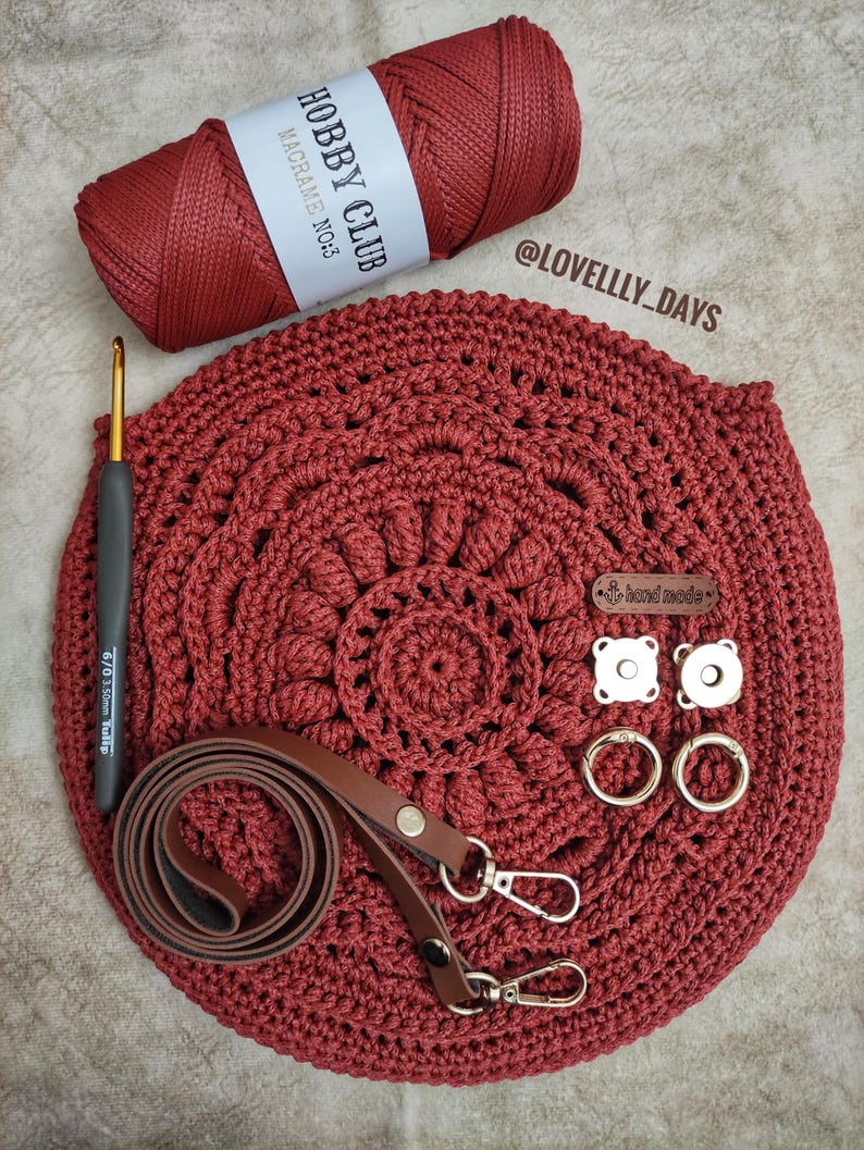 Pu&ograve; includere: Una borsa a uncinetto rossa con una tracolla in pelle marrone e accessori dorati. La borsa &egrave; realizzata con un cordoncino rosso etichettato "Hobby Club Macrame No.3". La borsa ha anche un piccolo cartellino "fatto a mano" e due chiusure magnetiche.