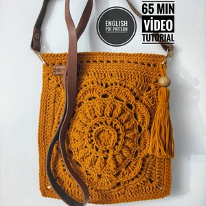 May include: A crocheted mustard yellow crossbody bag with a brown leather strap and a tassel. The bag features a circular design with a flower in the center. The text "ENGLISH PDF PATTERN" is visible in a circle on the bag. The text "PATTERN 65 MIN VIDEO TUTORIAL" is visible in a rectangle at the top of the image.