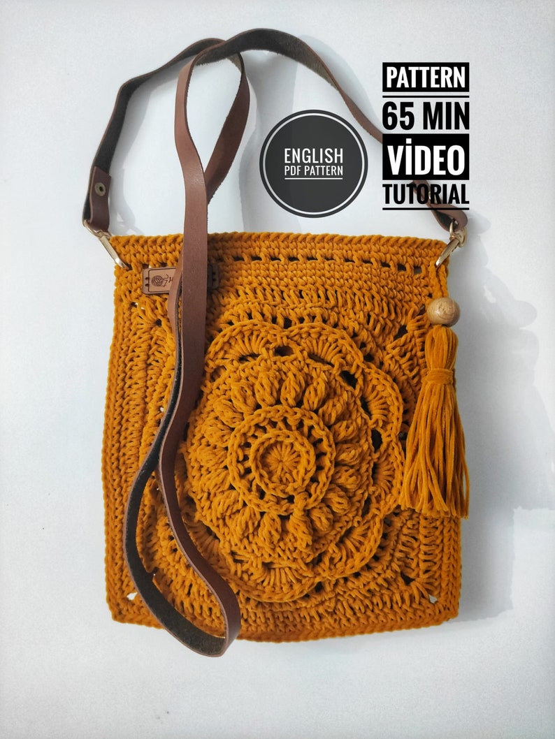 May include: A crocheted mustard yellow crossbody bag with a brown leather strap and a tassel. The bag features a circular design with a flower in the center. The text "ENGLISH PDF PATTERN" is visible in a circle on the bag. The text "PATTERN 65 MIN VIDEO TUTORIAL" is visible in a rectangle at the top of the image.