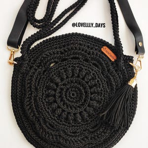 Puede incluir: Bolso redondo de crochet negro con un borla negra y una correa de cuero negro. El bolso tiene un cierre dorado y una peque&ntilde;a etiqueta de cuero marr&oacute;n con el texto "@LOVELLY_DAYS" en ella.