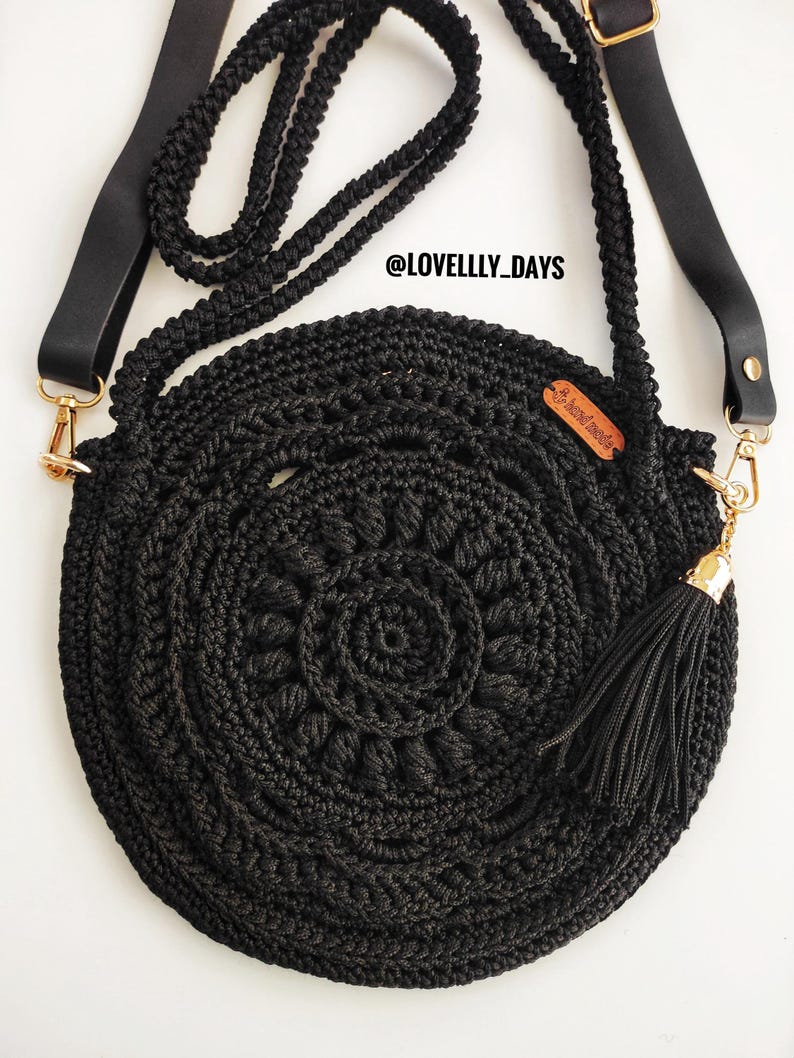 Puede incluir: Bolso redondo de crochet negro con un borla negra y una correa de cuero negro. El bolso tiene un cierre dorado y una peque&ntilde;a etiqueta de cuero marr&oacute;n con el texto "@LOVELLY_DAYS" en ella.