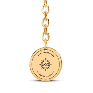 Op de afbeelding: Gouden sleutelhanger met een ronde hanger met een kompas en bergen met de tekst "Sint Christoffel, beschermheilige van reizigers".