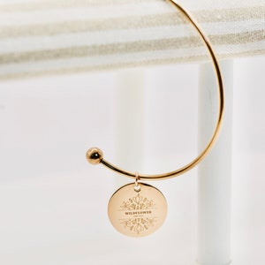 May include: Gold bangle bracelet with a round charm that says "Wildflower" with a floral design.