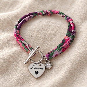 May include: A bracelet featuring a fabric band with a colorful floral pattern in shades of pink, purple, and green. It includes a silver-toned toggle clasp, a heart-shaped charm engraved with the name Louane and a small heart, and a crystal charm.