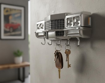 May include: Silver car front key holder mounted on a gray wall. The key holder has a grill, headlights, and hooks for keys. Several keys hang from the hooks. A framed picture and a wooden bench are in the background.