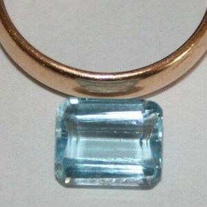 May include: A light blue aquamarine gemstone with an emerald cut. The gemstone is sitting on a white surface.