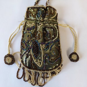 Könnte beinhalten: Eine kleine, perlenbesetzte Kordeltasche mit einer gold- und braunen Stoffbasis. Die Tasche hat unten einen Fransen aus Perlen und an jeder Seite einen Perlenquaste.