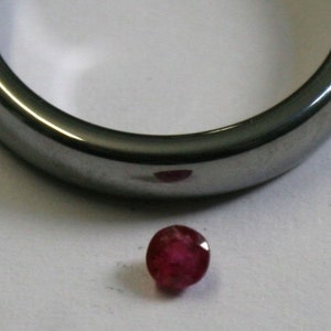 May include: A close-up of a small, round, red gemstone. The gemstone is sitting on a white surface.