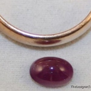 Può includere: Una pietra preziosa ovale di colore viola scuro, probabilmente un rubino, è mostrata su una superficie bianca. La pietra preziosa ha un diametro di circa 1 cm.