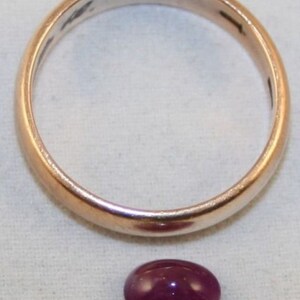 May include: A close-up of a deep purple oval gemstone. The gemstone is sitting on a white surface next to a gold band.
