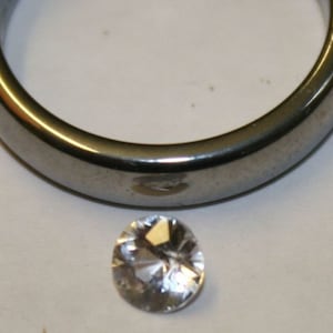 May include: A clear, round gemstone with facets, set against a silver metal background.