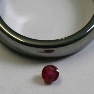 Può includere: Una pietra preziosa rotonda di colore rosso scuro con taglio a faccette. La pietra preziosa è su una superficie bianca.